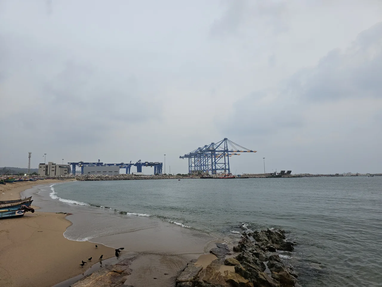Vizhinjam port cranes across the water, viewed from a nearby beach.